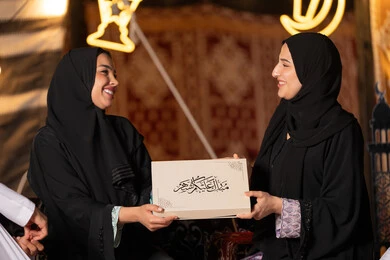 سيدتان سعوديتان تمسكان علبة هدايا عليها مخطوطة مبارك عليكم الشهر مع هلال وفوانيس رمضان باللون الاسود، التهنئه وتقديم الهديه في شهر رمضان المبارك، مناسبه دينيه إسلاميه، سيده عربيه خليجيه اماراتيه تقدم هديه لصديقتها