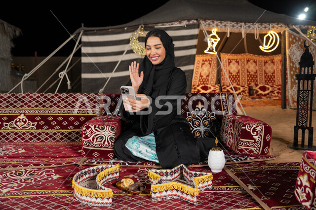 إيماءه الاشاره والترحيب، استخدام الهاتف للدردشه مع الأهل والأصدقاء عبر الانترنت، استخدام التكنولوجيا والتقنيات الحديثة، امرأة عربية سعودية خليجية محجبة تنظرالي الهاتف، سيده اماراتيه تقوم بمكالمه فيديو، استخدام التقنيات والاجهزه الحديثه