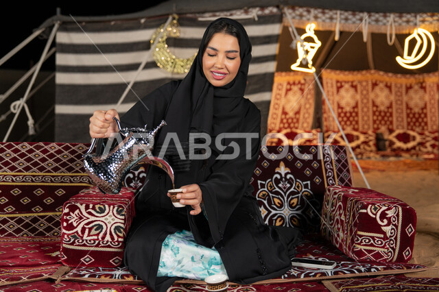 قيم وعادات الضيافه العربيه، حسن الاستقبال وكرم الضيافه،  سيده اماراتيه ترتدي العباءه السوداء تقوم بصب القهوه، امرأه عربيه خليجيه سعوديه مبتسمه تحمل الدله النحاسيه وكوب القهوه, المجلس الشعبي التقليدي