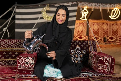 Values and customs of Arab hospitality, good reception and hospitality, an Emirati woman wearing a black abaya pouring coffee, a smiling Saudi Arabian Gulf woman carrying a copper coffee pot and a cup of coffee, the traditional folk council
