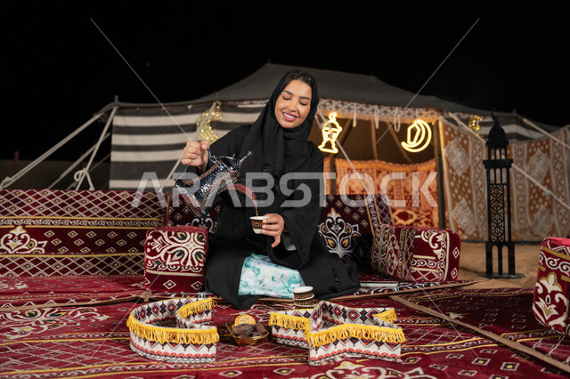 قيم وعادات الضيافه العربيه، حسن الاستقبال وكرم الضيافه،  سيده اماراتيه ترتدي العباءه السوداء تقوم بصب القهوه، امرأه عربيه خليجيه سعوديه مبتسمه تحمل الدله النحاسيه وكوب القهوه, المجلس الشعبي التقليدي