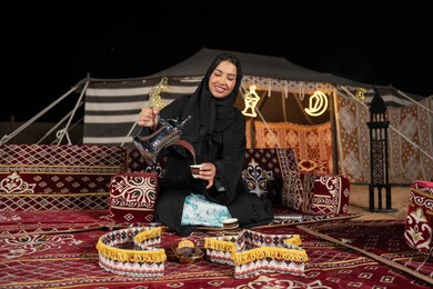 قيم وعادات الضيافه العربيه، حسن الاستقبال وكرم الضيافه،  سيده اماراتيه ترتدي العباءه السوداء تقوم بصب القهوه، امرأه عربيه خليجيه سعوديه مبتسمه تحمل الدله النحاسيه وكوب القهوه, المجلس الشعبي التقليدي