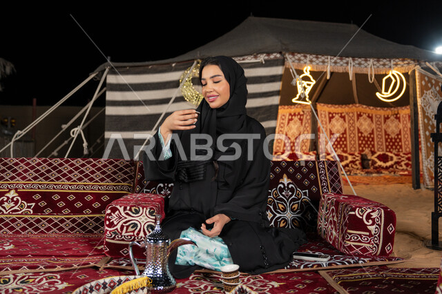 الاستمتاع بتناول القهوه العربيه الساخنه، سيده اماراتيه ترتدي العباءه السوداء تجلس في خيمه وقت الافطار, المجلس الشعبي التقليدي، امرأه عربيه خليجيه سعوديه تمسك كوب القهوه، حسن الاستقبال وكرم الضيافه عند العرب