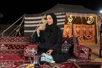 الاستمتاع بتناول القهوه العربيه الساخنه، سيده اماراتيه ترتدي العباءه السوداء تجلس في خيمه وقت الافطار, المجلس الشعبي التقليدي، امرأه عربيه خليجيه سعوديه تمسك كوب القهوه، حسن الاستقبال وكرم الضيافه عند العرب