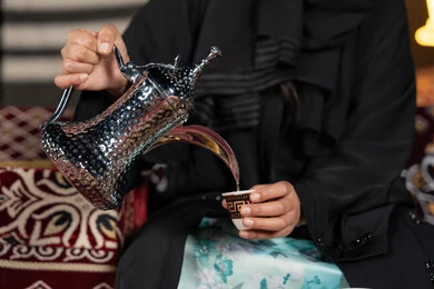 حسن الاستقبال وكرم الضيافه عند العرب،  سيده اماراتيه ترتدي العباءه السوداء تقوم بصب القهوه, المجلس الشعبي التقليدي، صوره مقربه ليدين امرأه عربيه خليجيه سعوديه تحمل الدله النحاسيه وكوب القهوه الساخنه ، ديكورات شهر رمضان الفضيل