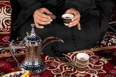 كسر الصيام بتناول حبه التمربجانب القهوه، سيده اماراتيه ترتدي العباءه السوداء تجلس في خيمه وقت الافطار, المجلس الشعبي التقليدي، صوره مقربه ليدين امرأه عربيه خليجيه سعوديه تمسك حبه التمر وكوب القهوه، حسن الاستقبال وكرم الضيافه عند العرب