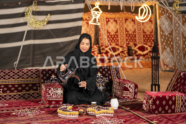قيم وعادات الضيافه العربيه، حسن الاستقبال وكرم الضيافه،  سيده اماراتيه ترتدي العباءه السوداء تقوم بصب القهوه، امرأه عربيه خليجيه سعوديه مبتسمه تحمل الدله النحاسيه وكوب القهوه, المجلس الشعبي التقليدي