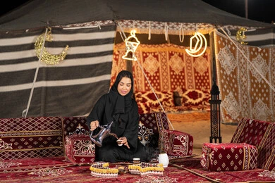 قيم وعادات الضيافه العربيه، حسن الاستقبال وكرم الضيافه،  سيده اماراتيه ترتدي العباءه السوداء تقوم بصب القهوه، امرأه عربيه خليجيه سعوديه مبتسمه تحمل الدله النحاسيه وكوب القهوه, المجلس الشعبي التقليدي