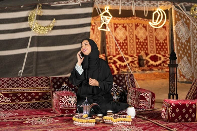 اجراء مكالمه هاتفيه مع شخص ما، سيده اماراتيه ترتدي العباءه السوداء تقوم باجراء مكالمه مع الاصدقاء، امرأه عربيه خليجيه سعوديه مبتسمه تقوم باستخدام الهاتف المحمول, المجلس الشعبي التقليدي، التقدم التقني للاجهزه، التواصل عن بعد، ديكورات شهر رمضان المبارك