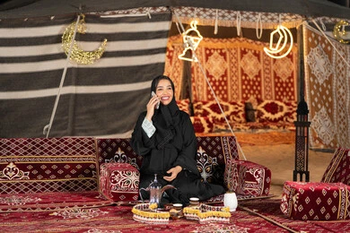 اجراء مكالمه هاتفيه مع شخص ما، سيده اماراتيه ترتدي العباءه السوداء تقوم باجراء مكالمه مع الاصدقاء، امرأه عربيه خليجيه سعوديه مبتسمه تقوم باستخدام الهاتف المحمول, المجلس الشعبي التقليدي، التقدم التقني للاجهزه، التواصل عن بعد، ديكورات شهر رمضان المبارك