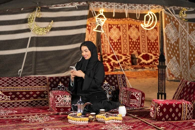 Chatting, texting, and remote communication. An Emirati woman wearing a black abaya is making a video call with friends. A smiling Saudi Arabian Gulf woman is using a mobile phone. Traditional popular council. Technical progress of devices. Communication via the Internet.