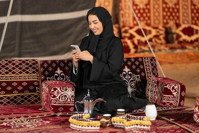 Chatting, texting, and remote communication. An Emirati woman wearing a black abaya is making a video call with friends. A smiling Saudi Arabian Gulf woman is using a mobile phone. Traditional popular council. Technical progress of devices. Communication via the Internet.