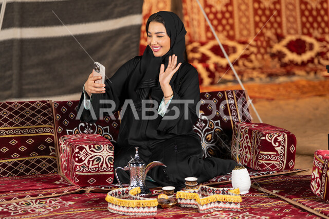 الاشاره والترحيب لشخص ما، سيده اماراتيه ترتدي العباءه السوداء تقوم باجراء مكالمه فيديو مع الاصدقاء، امرأه عربيه خليجيه سعوديه مبتسمه تقوم باستخدام الهاتف المحمول, المجلس الشعبي التقليدي، التقدم التقني للاجهزه، التواصل عبرالانترنت