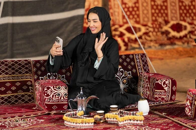الاشاره والترحيب لشخص ما، سيده اماراتيه ترتدي العباءه السوداء تقوم باجراء مكالمه فيديو مع الاصدقاء، امرأه عربيه خليجيه سعوديه مبتسمه تقوم باستخدام الهاتف المحمول, المجلس الشعبي التقليدي، التقدم التقني للاجهزه، التواصل عبرالانترنت