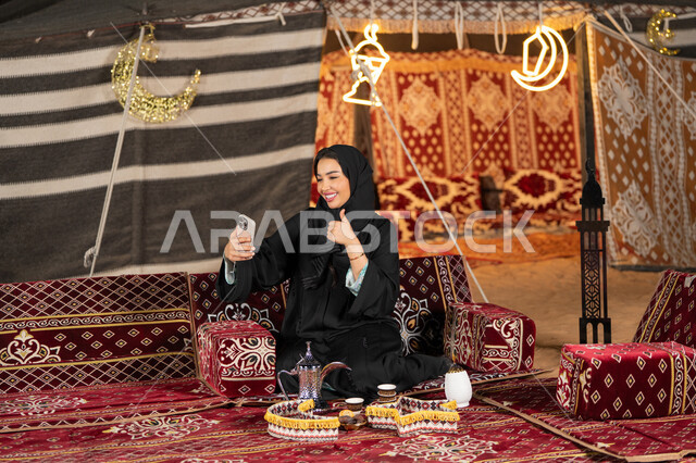ديكور وزينه شهر رمضان، سيده اماراتيه ترتدي العباءه السوداء تقوم باجراء مكالمه فيديو مع الاصدقاء، امرأه عربيه خليجيه سعوديه مبتسمه تقوم باستخدام الهاتف المحمول, المجلس الشعبي التقليدي، التقدم التقني للاجهزه، التواصل عن بعد، ايماءه الاعجاب والسرور
