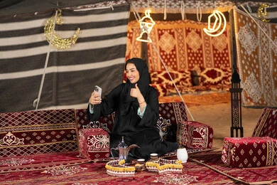 ديكور وزينه شهر رمضان، سيده اماراتيه ترتدي العباءه السوداء تقوم باجراء مكالمه فيديو مع الاصدقاء، امرأه عربيه خليجيه سعوديه مبتسمه تقوم باستخدام الهاتف المحمول, المجلس الشعبي التقليدي، التقدم التقني للاجهزه، التواصل عن بعد، ايماءه الاعجاب والسرور