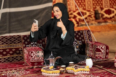 Emirati woman wearing black abaya making a video call with friends, smiling Saudi Arabian Gulf woman using mobile phone, night camping, traditional folk council, technical advancement of devices, remote communication, gesture of admiration and pleasure