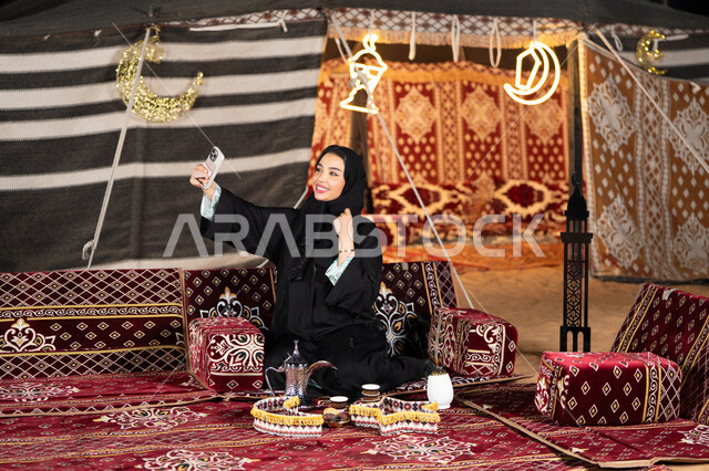 التقاط صور سيلفي تذكاريه، سيده اماراتيه ترتدي العباءه السوداء تمسك بيديها جهاز الهاتف، امرأه عربيه خليجيه سعوديه تجلس في خيمه مليئه بزينه وديكورات شهر رمضان, التخييم الليلي، المجلس الشعبي التقليدي، كرم الضيافه وحسن الاستقبال، التقدم التقني للاجهزه