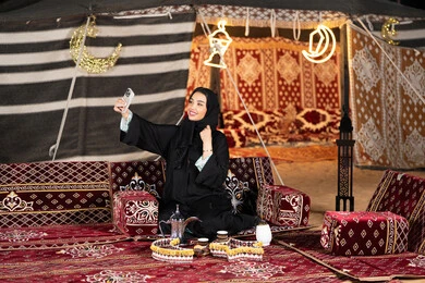 التقاط صور سيلفي تذكاريه، سيده اماراتيه ترتدي العباءه السوداء تمسك بيديها جهاز الهاتف، امرأه عربيه خليجيه سعوديه تجلس في خيمه مليئه بزينه وديكورات شهر رمضان, التخييم الليلي، المجلس الشعبي التقليدي، كرم الضيافه وحسن الاستقبال، التقدم التقني للاجهزه