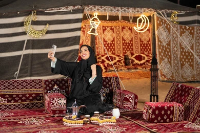 Taking souvenir selfies, an Emirati woman wearing a black abaya holding a phone in her hands, a Saudi Arabian Gulf woman sitting in a tent filled with Ramadan decorations, night camping, traditional Majlis, generous hospitality and warm reception, technical advancement of devices