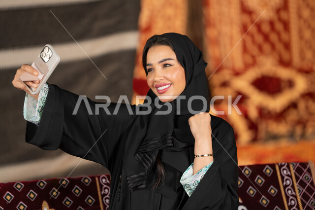 توثيق اللحظات السعيده بالتقاط الصور التذكاريه، سيده اماراتيه ترتدي العباءه السوداء تمسك بيديها جهاز الهاتف، امرأه عربيه خليجيه سعوديه مبتسمه تقوم بالتقاط صوره سيلفي, التخييم الليلي، المجلس الشعبي التقليدي، التقدم التقني للاجهزه