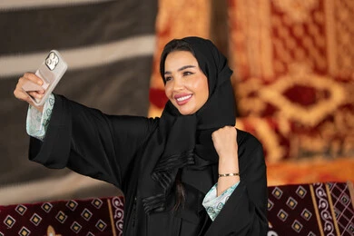 توثيق اللحظات السعيده بالتقاط الصور التذكاريه، سيده اماراتيه ترتدي العباءه السوداء تمسك بيديها جهاز الهاتف، امرأه عربيه خليجيه سعوديه مبتسمه تقوم بالتقاط صوره سيلفي, التخييم الليلي، المجلس الشعبي التقليدي، التقدم التقني للاجهزه
