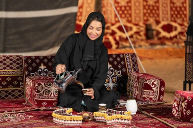 Inherited principles of serving Arabic coffee, an Emirati woman wearing a black abaya holding a copper coffee pot and a cup of coffee, a Saudi Arabian Gulf woman sitting in a tent serving hot coffee, night camping, the traditional popular council, generous hospitality and warm welcome