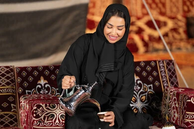 اصول متوارثه لتقديم القهوه العربيه، سيده اماراتيه ترتدي العباءه السوداء تمسك بيديها دله نحاسيه وكوب من القهوه، امرأه عربيه خليجيه سعوديه تجلس في خيمه تقوم بتقديم القهوه الساخنه, التخييم الليلي، المجلس الشعبي التقليدي، كرم الضيافه وحسن الاستقبال