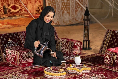 توارث العادات من الاجداد لتقديم القهوه، سيده اماراتيه ترتدي العباءه السوداء تمسك بيديها دله نحاسيه وكوب من القهوه، امرأه عربيه خليجيه سعوديه تجلس في خيمه تقوم بتقديم القهوه الساخنه, التخييم الليلي، المجلس الشعبي التقليدي، كرم الضيافه وحسن الاستقبال