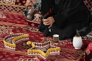 Emirati woman wearing black abaya holding a copper coffee pot and a cup of coffee, close-up of the hands of a Saudi Arabian Gulf woman sitting in a tent in the middle of the deserts of the Kingdom serving hot coffee, quiet night camping, traditional folk council, generous hospitality and warm welcome