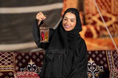 خيمه مليئه بزينه وديكورات رمضانيه, سيده اماراتيه ترتدي العباءه والحجاب الاسود تمسك بفانوس معدني ملون، امرأه عربيه خليجيه سعوديه مبتسمه تحتفل بقدوم شهر رمضان المبارك, التخييم الليلي الهادئ، المجلس الشعبي التقليدي