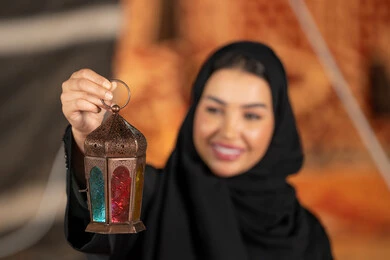 سيده اماراتيه ترتدي العباءه والحجاب الاسود تمسك بفانوس معدني ملون، صوره مقربه ليد امرأه عربيه خليجيه سعوديه مبتسمه تحتفل بقدوم شهر رمضان المبارك, خيمه مليئه بزينه وديكورات رمضانيه, التخييم الليلي الهادئ