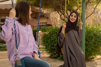 نزهة في الهواء الطلق، بناء علاقات اجتماعية ناجحة  ،تبادل أطراف الحديث بين العائلات والزملاء، الاستمتاع بعطلة نهاية الأسبوع برفقة الأصدقاء في المطاعم والكافيهات، صديقتان  عربيتان خليجيتان سعوديتان تجلسان على المرجيحة باحدي الحدائق الترفهيه بالسعودية ،يتبادلان الاحاديث في الأجواء الصيفية اللطيفة