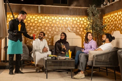 تبادل أطراف الحديث بين العائلات والزملاء، نادل يرتدي زي اليونيفورم يقوم بتقديم الطلبات ،  الاستمتاع بعطلة نهاية الأسبوع برفقة الأصدقاء في المطاعم والكافيهات،مجموعة اصدقاء عرب خليجيين سعوديين يقضون السهرات المسائية الممتعة في الأجواء الصيفية اللطيفة ، تناول المشروبات الباردة المنعشة  في الهواء الطلق