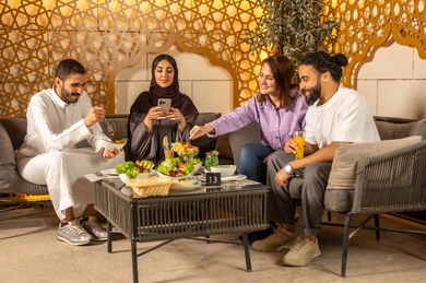 A group of Arab Gulf friends from Saudi Arabia spend enjoyable evening gatherings in the pleasant summer atmosphere, using mobile phones, exchanging conversations between families and colleagues, enjoying the weekend with friends at restaurants and cafes, and having refreshing cold drinks outdoors.