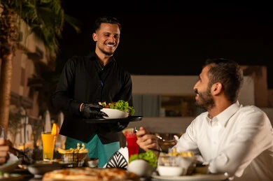 Pleasant summer atmosphere, enjoying cold and refreshing seasonal juices, working in luxurious restaurants and cafes in Saudi Arabia, a waiter in a uniform serving orders to customers, an Arab Gulf Saudi man enjoying his time in an outdoor seating area of the restaurant.