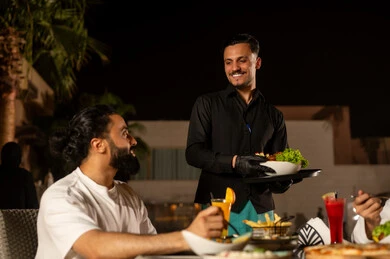 A waiter in a uniform is serving orders to customers, working in luxury restaurants and cafes in Saudi Arabia, enjoying cold and refreshing seasonal juices, an Arab Gulf Saudi man is enjoying his time in an outdoor seating area of the restaurant in a pleasant summer atmosphere.