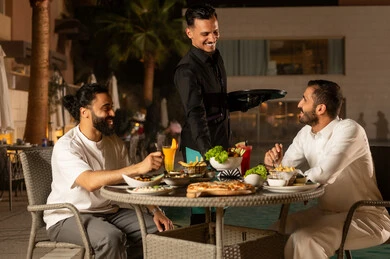 تناول العصائر الموسمية الباردة والمنعشة، العمل في المطاعم والكافيهات الفاخرة في السعودية، نادل يرتدي زي اليونيفورم يقوم بتقديم الطلبات للزبائن، صديقان عربيان خليجيان سعوديان يستمتعان بوقتهما في جلسة خارجية للمطعم ضمن أجواء صيفية لطيفة