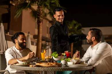 Working in luxury restaurants and cafes in Saudi Arabia, enjoying cold and refreshing seasonal juices, a waiter in a uniform serving orders to customers, two Arab Gulf friends from Saudi Arabia enjoying their time in an outdoor seating area of the restaurant in a pleasant summer atmosphere.