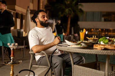 A fun and entertaining gathering in pleasant summer weather, engaging in enjoyable activities during the weekend, smoking shisha and having cold drinks, an Arab Gulf Saudi man sitting outdoors, visiting restaurants and cafes in the Kingdom of Saudi Arabia.