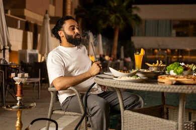 Smoking shisha and drinking cold beverages, engaging in fun activities on the weekend, a relaxing and entertaining session in pleasant summer weather, an Arab Gulf Saudi man sitting outdoors, visiting restaurants and cafes in the Kingdom of Saudi Arabia.
