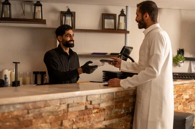 استخدام التقنيات الحديثة ، أسلوب الضيافة الراقية في المقاهي السعودية ، الدفع الالكتروني عبر بطاقة الائتمان ،نادل مبتسم يقدم الماكينة لرجل سعودي يرتدي الزي السعودي التقليدي  وهو  يستخدم هاتفه للدفع ، احدي الكافيهات الحديثة في السعودية
