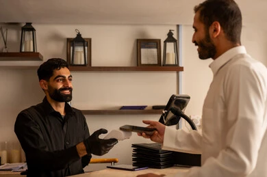 الخروج فى  احدي الكافيهات الحديثة في السعودية، أسلوب الضيافة الراقية في المقاهي السعودية ، الدفع الالكتروني عبر بطاقة الائتمان ،نادل مبتسم يقدم الماكينة لرجل سعودي يرتدي الزي السعودي التقليدي  وهو  يستخدم هاتفه للدفع