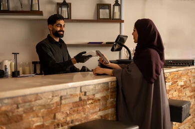 A smiling waiter presents the machine to a Saudi woman wearing an abaya and hijab as she uses her phone to pay. This is a reflection of the high-end hospitality style in Saudi cafes, with electronic payment via credit card, in one of the modern cafes in Saudi Arabia.
