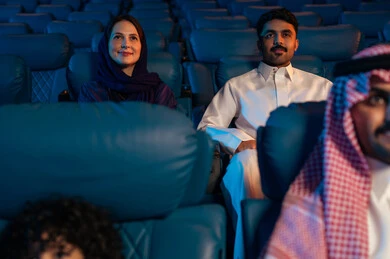 Modern display technologies, an Arab Saudi Gulf audience in a cinema hall, a four-dimensional screening room, advanced audio, visual, and sensory cinematic effects, a fantastic cinematic experience, enjoying popcorn, facial expressions indicating joy and happiness, a family entertainment activity.