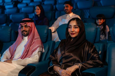 An Arab Saudi Gulf audience in a cinema hall, a four-dimensional screening room, modern display technologies, advanced audio, visual, and sensory cinematic effects, a fantastic cinematic experience, enjoying popcorn, facial expressions indicating joy and happiness, a family entertainment activity.