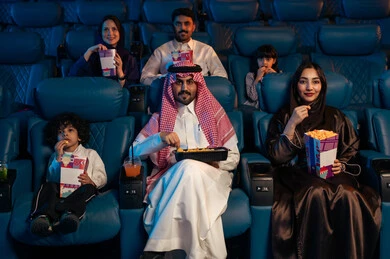 Modern display technologies, an Arab Saudi Gulf audience in a cinema hall, a four-dimensional screening room, advanced audio, visual, and sensory cinematic effects, a fantastic cinematic experience, enjoying popcorn, facial expressions indicating joy and happiness, a family entertainment activity.