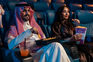 قضاء أوقات ممتعة، أنشطة ترفيهية سعيدة، رجل عربي سعودي خليجي بجانب زوجته في صالة السينما، تعابير تدل على الإندماج، الإستمتاع بتناول الفشار والمشروبات الباردة ، مشاهدة الأفلام والعروض المسرحية في السينما، سينما ومسرح، خلفية قاعة عرض السينما، صالة العرض