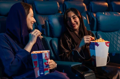 Enjoying popcorn, watching movies, plays, and various shows, large screens to enhance the experience, engaging in fun recreational activities, two Saudi Arabian Gulf friends sitting in a cinema hall watching the movie with gestures of happiness, modern theaters in the Kingdom of Saudi Arabia.
