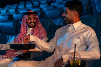 Watching movies, plays, and various shows, enjoying golden-colored snacks, large screens to enhance the experience, engaging in fun recreational activities, two Saudi Arabian Gulf men sitting in a cinema hall watching the movie with gestures of happiness, modern theaters in the Kingdom of Saudi Arabia.